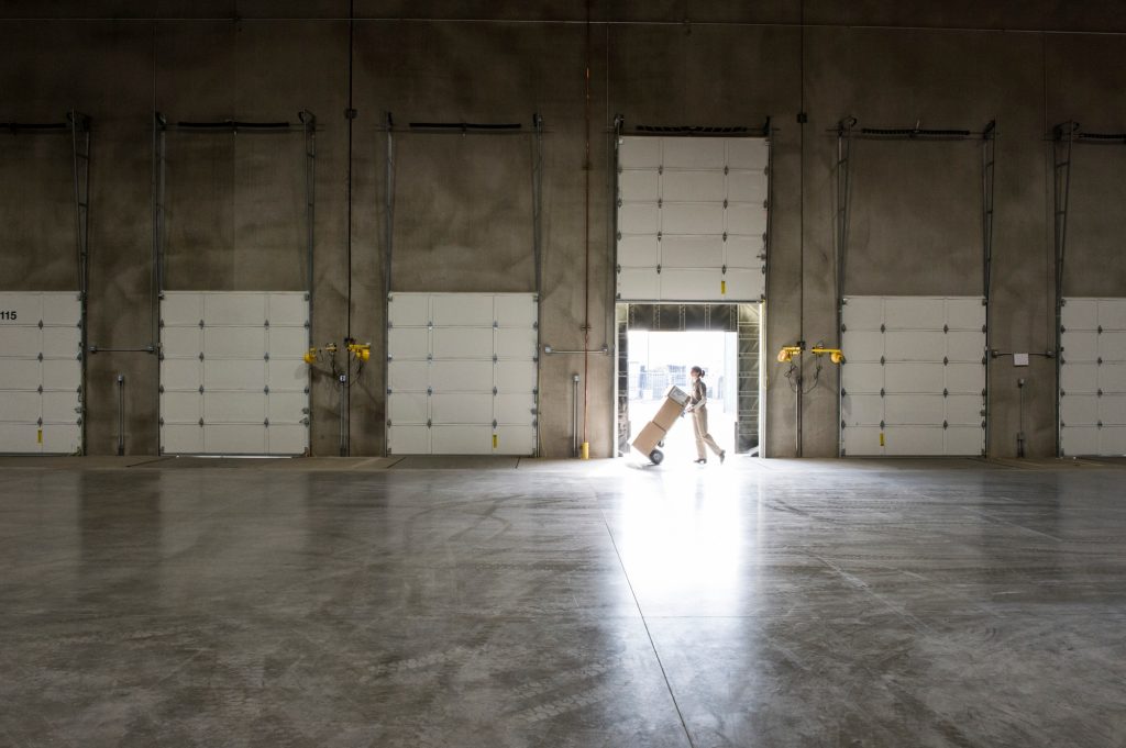 A warehouse worker with a hand truck at a loading dock door in a warehouse.